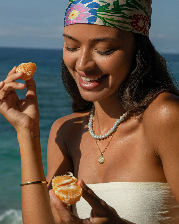 Colorful beaded necklace with a gold necklace by a woman holding tangerines