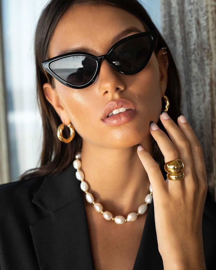 model close up wearing black sunglasses, pearl necklace, hoop earrings, and gold rings